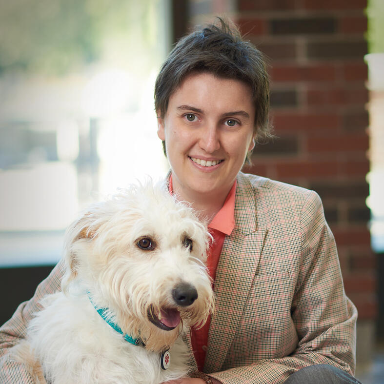 headshot of a white person with short brown hair wearing a coral shirt and blazer. They are posing with a scruffy white dog.